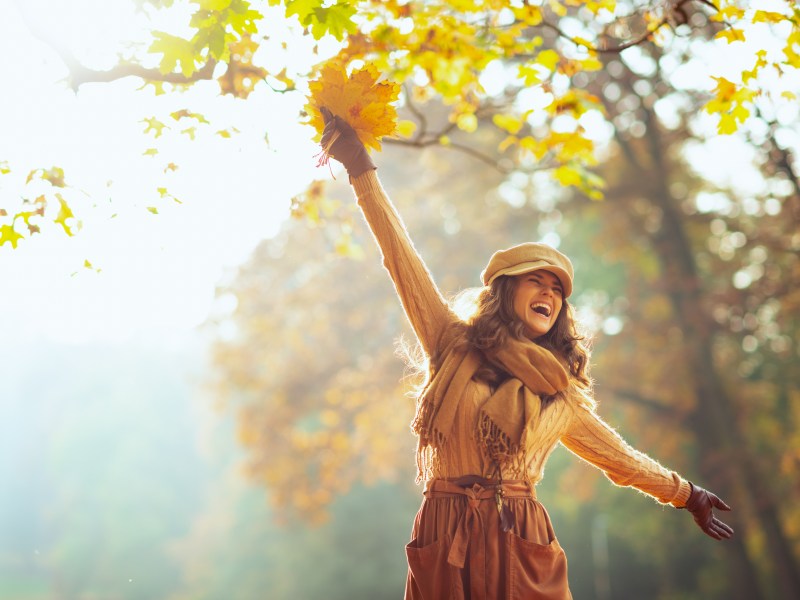 Frau im Herbstoutfit, mit gelben Blättern in der Hand im herbstlichen Wald