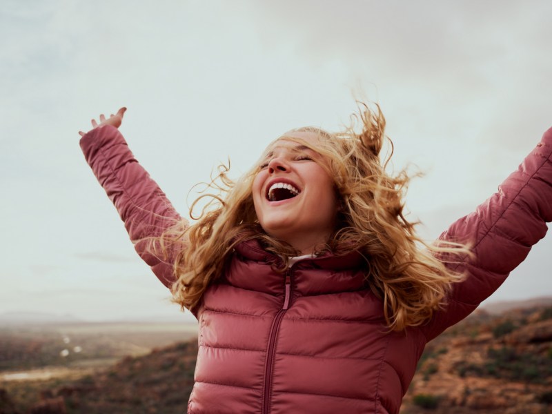 Frau mit rosa WInterjacke, die die Arme in die Luft schwingt und lacht, während sie draußen auf einem Berg steht