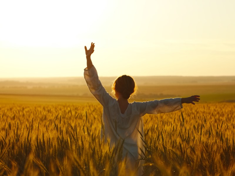 Frau in einem Feld im Sonnenuntergang, die ihre Arme in die Luft hält