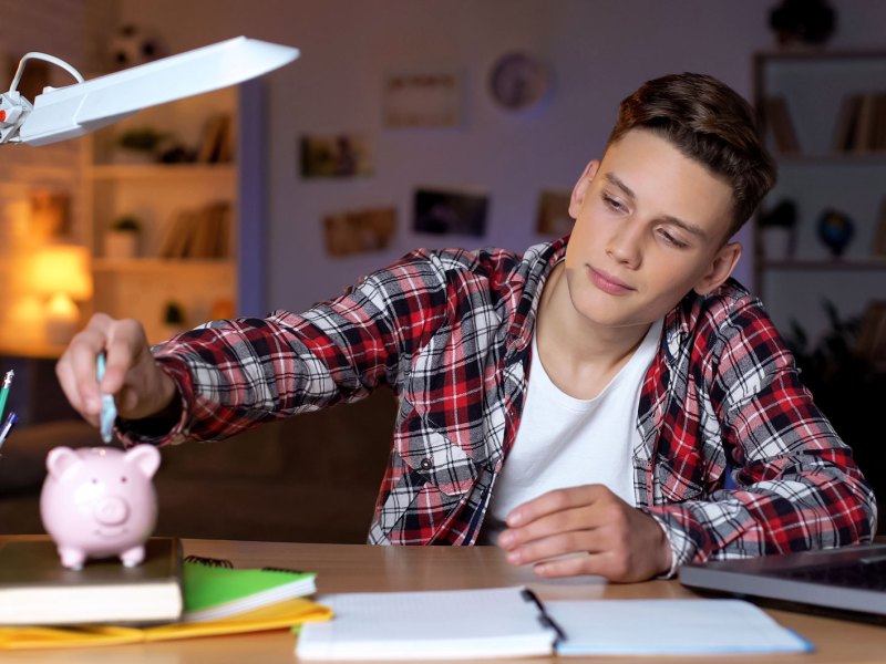 Teenager sitzt an seinem Schreibtisch und schiebt einen Geldschein in sein Sparschwein.