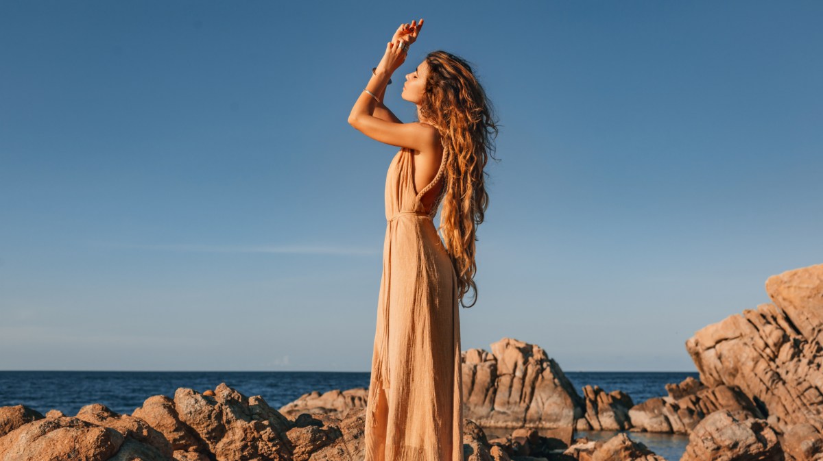 Frau auf Steinen vor einer Bucht, die ein langes Kleid anhat und ihre Hände in die Luft streckt