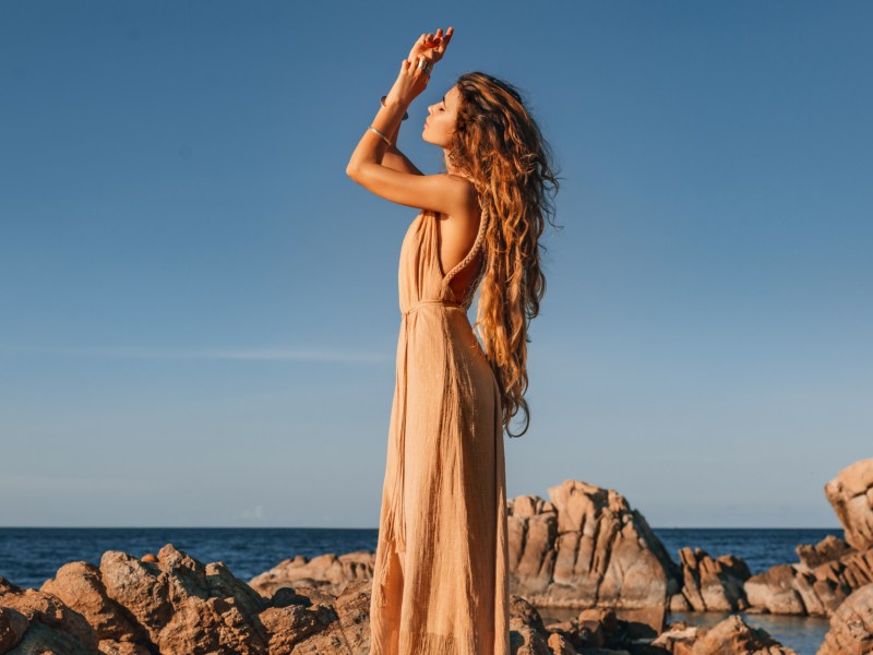 Frau auf Steinen vor einer Bucht, die ein langes Kleid anhat und ihre Hände in die Luft streckt