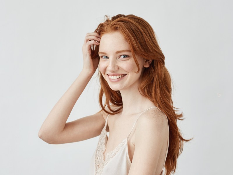 Junge Frau mit roten Haare, die schüchtern zur Seite guckt und vor einer weißen Wand steht
