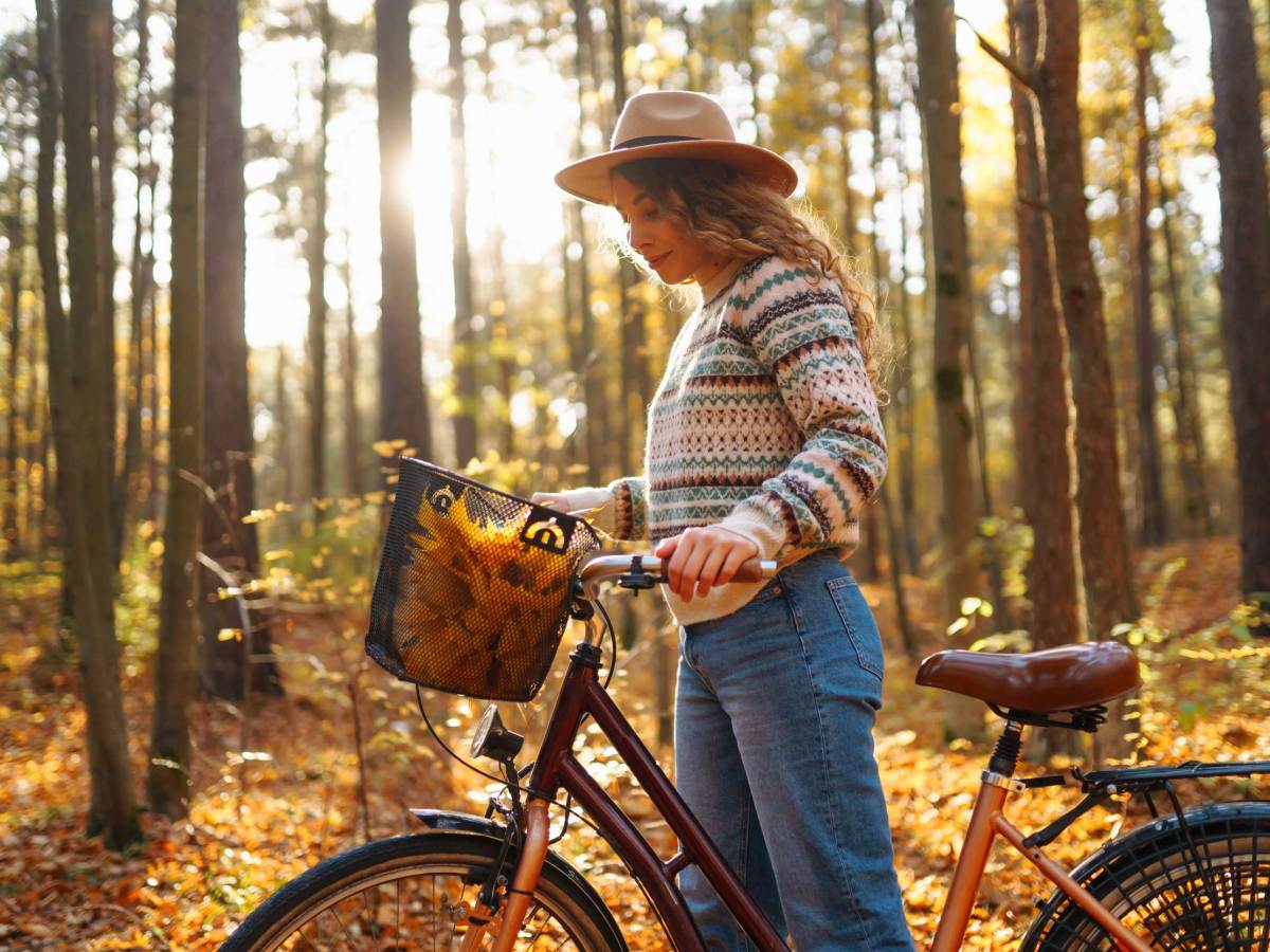 Lächelnde junge Frau in einem Hut und einem stilvollen Pullover und einem Fahrrad, genießt das Herbstwetter im Wald, unter den gelben Blättern.