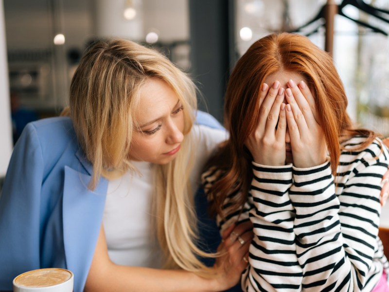 Zwei Frauen in einem Café, die nebeneinander sitzen, während die linke, blonde Frau versucht, die rothaarige, rechte Frau zu trösten