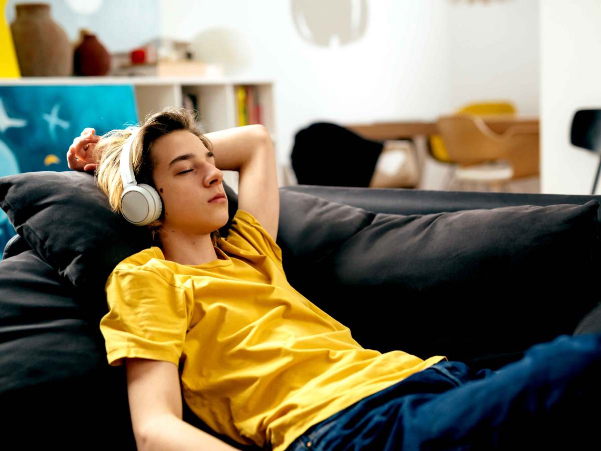 Männlicher Teenager liegt auf dem Sofa, trägt Kopfhörer und hat die Augen geschlossen.