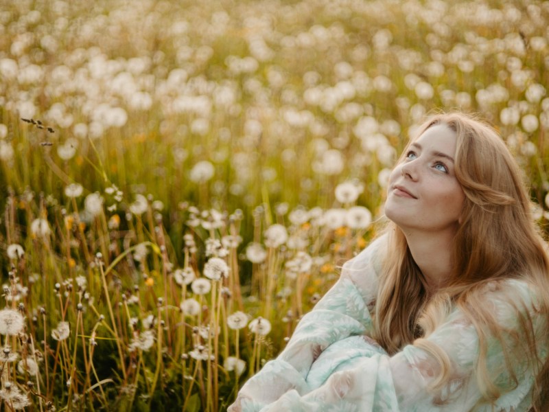 Frau in einem Feld voller Pusteblumen, die in den Himmel schaut und lächelt