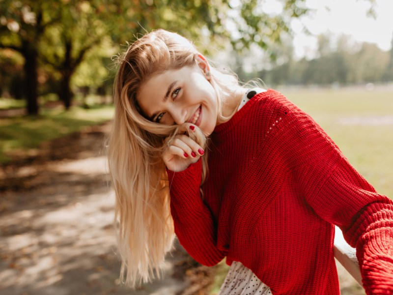 Frau im Park mit rotem Oberteil und blonden Haaren, die geschmeichelt lächelt