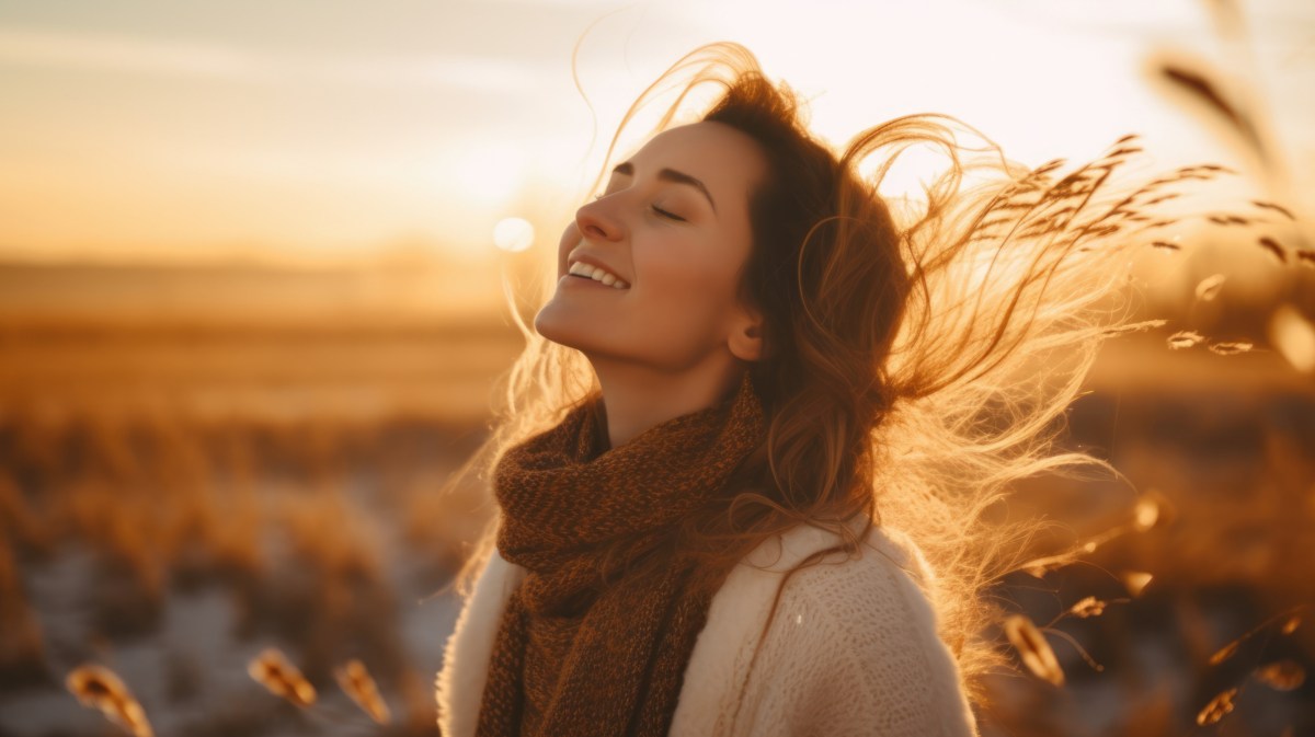 Frau mit geschlossenen Augen bei Sonnenuntergang auf einem Feld, lächelt. Haare wehen im Wind