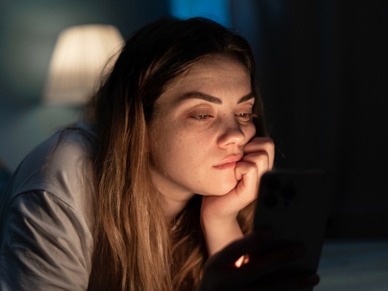 Frau, die gelangweilt in der Dunkelheit auf ihrem Bett liegt und auf ihr Handy Display starrt