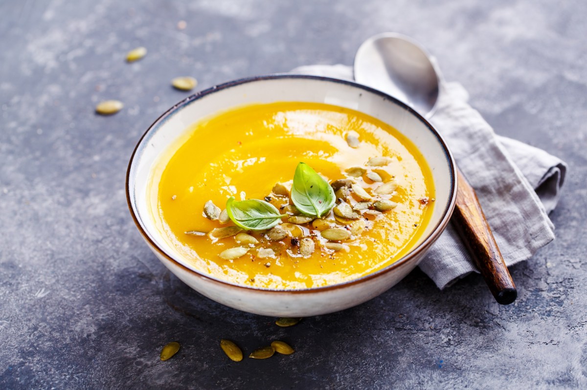 Eine weiße Schüssel Tomaten-Möhren-Suppe garniert mit Kürbiskernen und Basilikum, daneben ein Löffel mit Holzgriff auf einer Serviette.