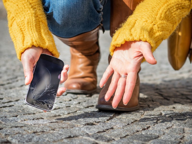 Frauenhände heben ein kaputtes Handy von einer Pflastersteinstraße auf.