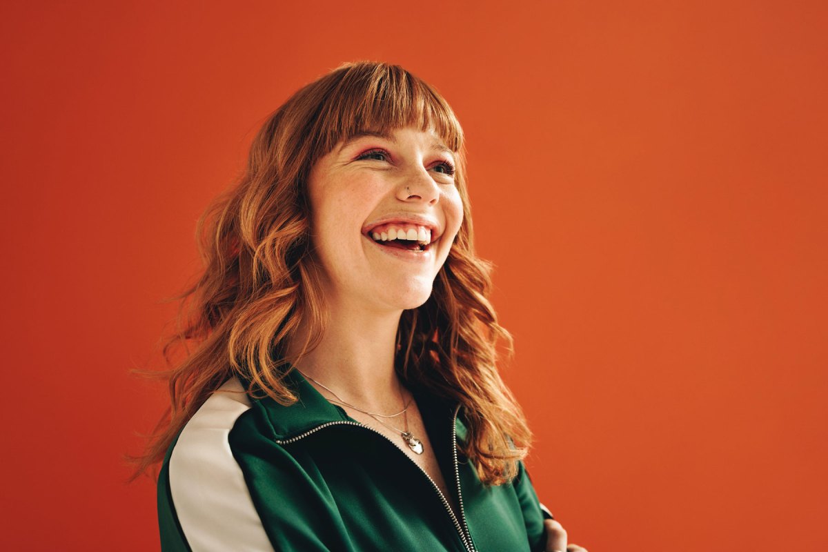 Eine Frau vor einer orangefarbenen Wand lacht.