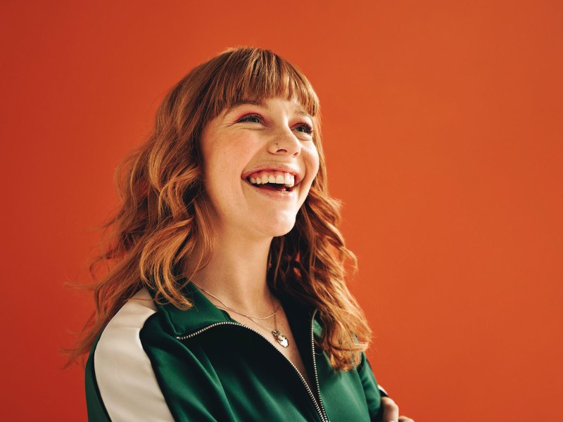Eine Frau vor einer orangefarbenen Wand lacht.