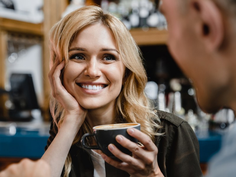 Blonde Frau hält eine Kaffeetasse in der Hand und sieht ihr Gegenüber verliebt an.