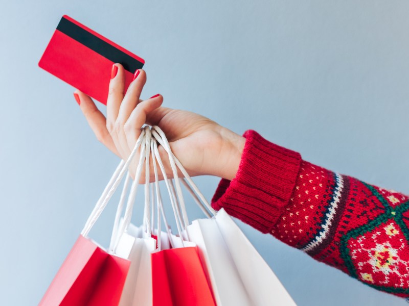 Frau mit Weihnachtspullover streckt ihren Arm raus und hält eine Kreditkarte und ein paar Tüten in der Hand.