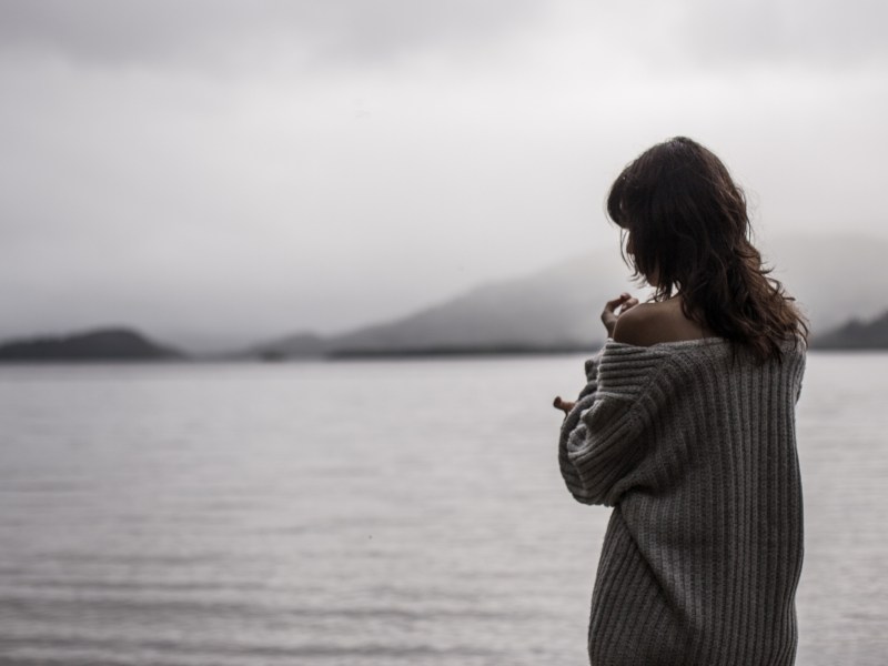 Frau mit grauem Pullover steht vor einem großen See.