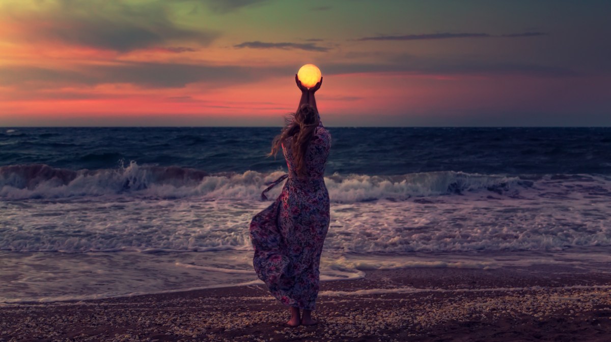 Frau mit einem Kleid und langen Haaren steht bei Sonnenuntergang am Strand und hält die Sonne in den Händen, wobei es sich um eine optische Täuschung handelt.