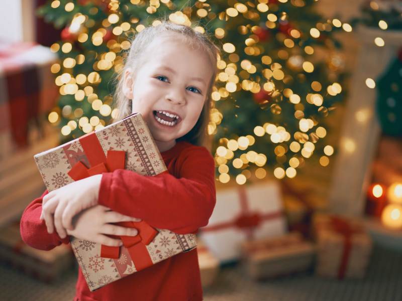 Kind hält ein Weihnachtsgeschenk fest im Arm und steht vor dem Tannenbaum.