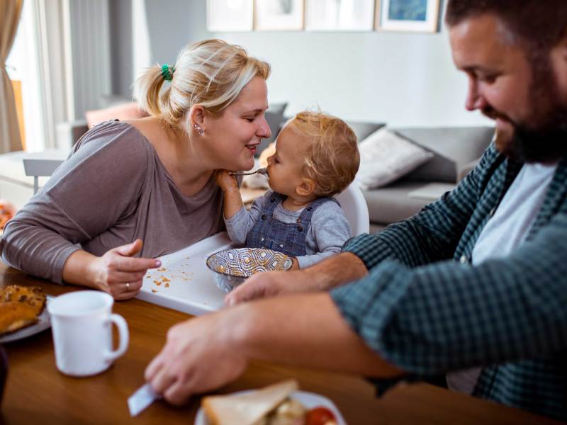 Vater, Mutter und Kleinkind sitzen gemeinsam am Frühstückstisch.
