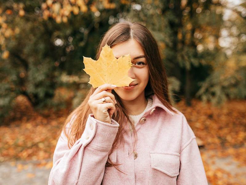 Positive braunhaarige Frau mit langen glatten Haaren in gemütlicher Herbstkleidung hat Spaß vor der Kamera, posiert mit Blatt auf ihrem Gesicht im Herbstpark. Altweibersommer.