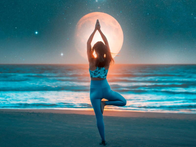 Frau macht Yoga-Übung am Strand, im Hintergrund ist der Mond