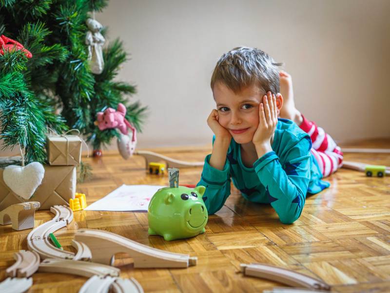 Kleiner Junge liegt neben dem Weihnachtsbaum, mit einem Sparschwein vor sich, in dem ein Geldschein steckt.