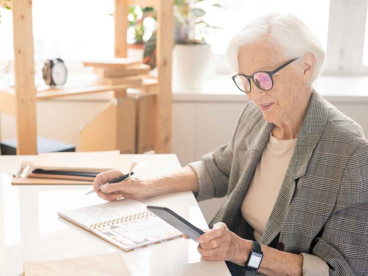 Ältere Frau sitzt am Tisch und schreibt etwas auf. Dabei hat sie ihr Handy in der Hand.