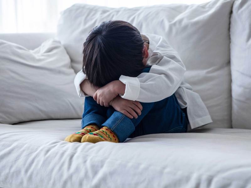Kleiner Junge sitzt auf dem Sofa und vergräbt seinen Kopf zwischen seinen Armen und Beinen.