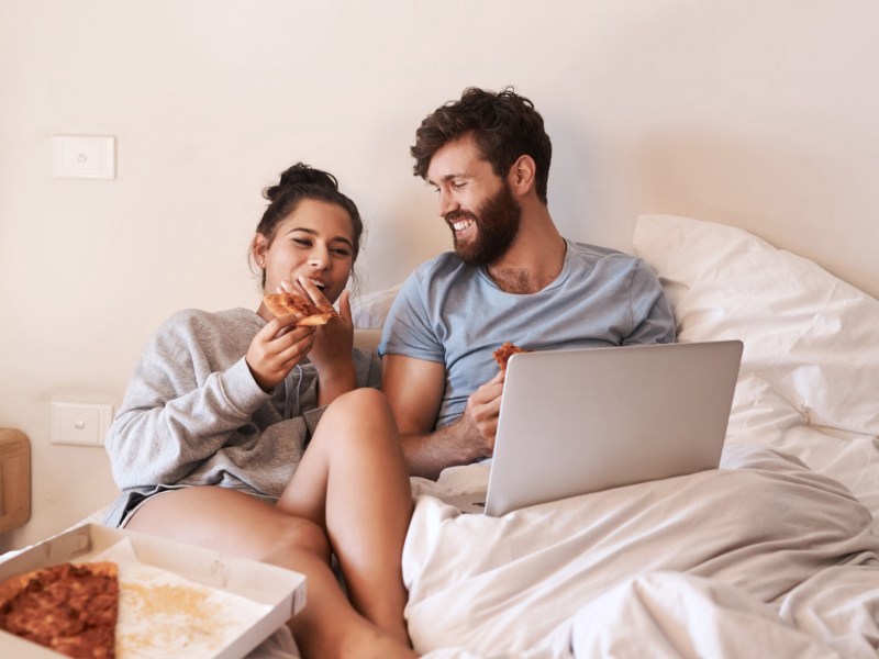 Mann und Frau im Bett, die einen Laptop auf der Bettdecke haben und Pizza essen