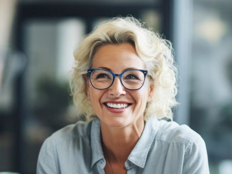 Frau mittleren Alters mit Brille und blonden kurzen Locken lächelt auf der Arbeit.