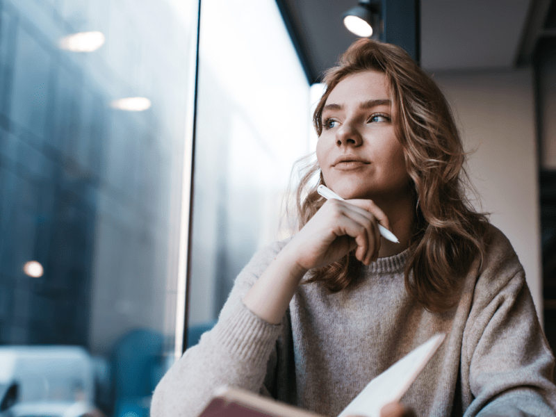 Frau vor einem Fenster in einem Café, die ein Buch in der Hand hält und einen Stift an ihr Kinn hält