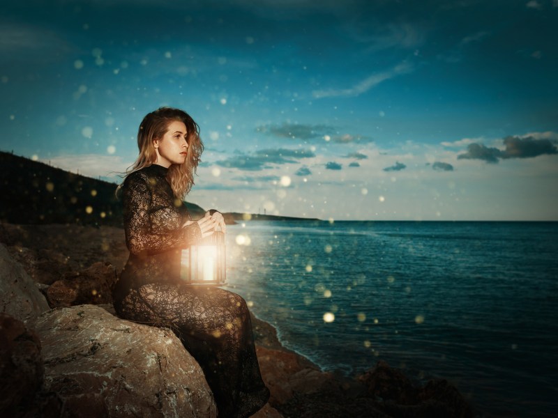 Frau sitzt mit einer Laterne an einem Felsen am Meer.