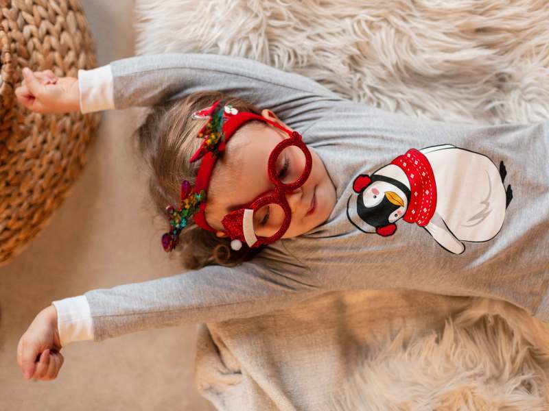 Kleines Mädchen Liegt in Weihnachtspulli und mit Partybrille auf einem Teppich und streckt sich vor Müdigkeit.