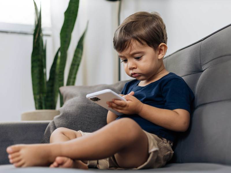 Kleiner, ca. 3-jähriger Junge sitzt mit einem Smartphone auf dem Sofa.
