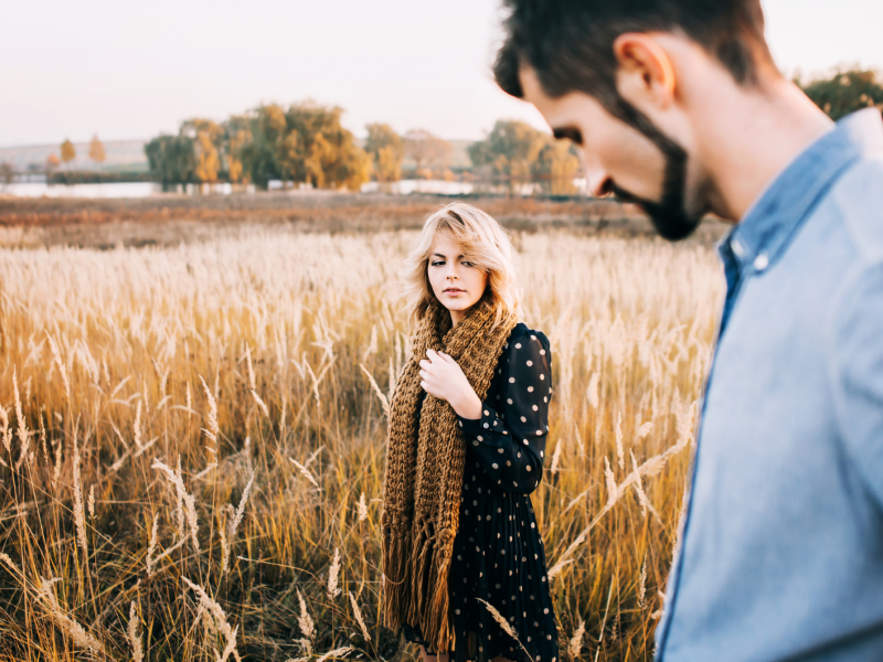 Frau mit lockigem Haar und Strickschal betrachtet nachdenklich eine weite, goldene Herbstlandschaft, ein Mann im Vordergrund unscharf.