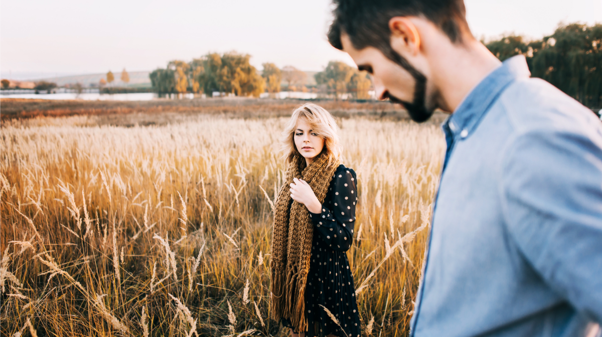 Frau mit lockigem Haar und Strickschal betrachtet nachdenklich eine weite, goldene Herbstlandschaft, ein Mann im Vordergrund unscharf.