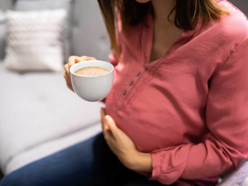Schwangere Frau sitzt auf dem Sofa, hat eine Hand auf ihren Bauch gelegt und hält in der anderen eine Tasse Kaffee.