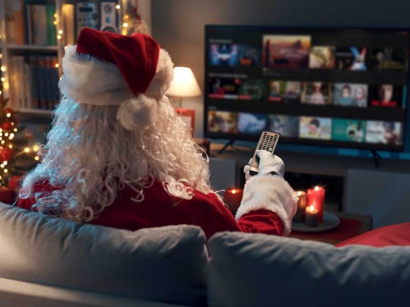 Weihnachtsmann sitzt mit dem Rücken zur Kamera auf der Couch vor einem Fernseher und überlegt, was er schauen kann. Neben ihm steht ein Weihnachtsbaum.