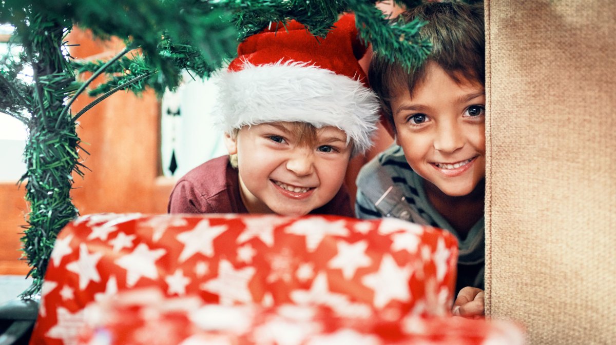 Zwei Jungs schauen neugierig unter dem Tannenbaum hindurch zu Geschenken.