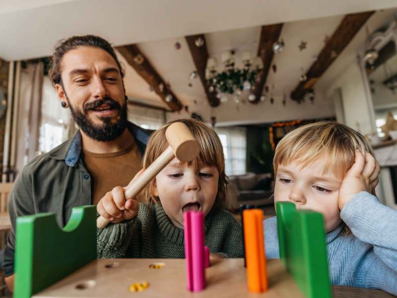 Vater spielt mit seinen beiden Kindern. Eines hat einen Holzhammer in der Hand und versucht, Holznägel in ein Brett zu hämmern.