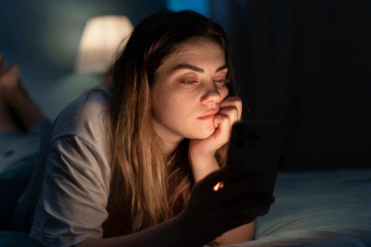 Eine Frau liegt im Bett und schaut gelangweilt auf ihr Handy.