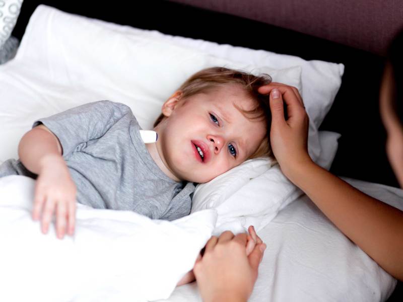 Kleine Junge liegt fiebrig im Bett, das Thermometer unter den Arm geklemmt. Seine Mutter sitzt neben dem Bett und hält seine Hand.