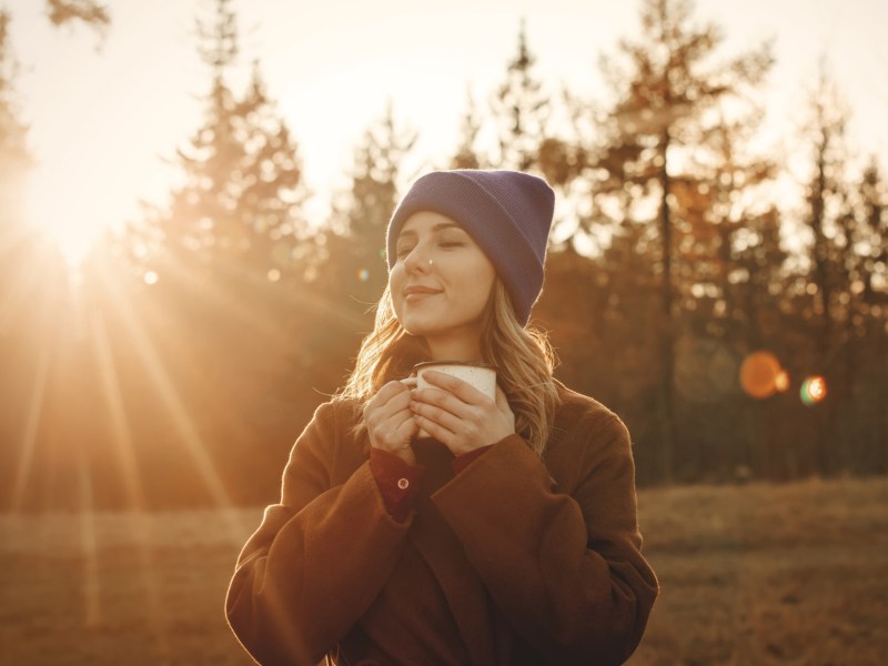 Frau im Wald mit einer Tasse in der Hand, die von Sonnenstrahlen angeleuchtet wird
