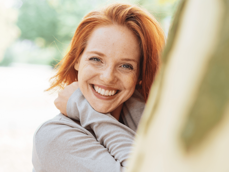 Frau mit roten Haaren vor einem Baumstamm, die in die Kamera lächelt
