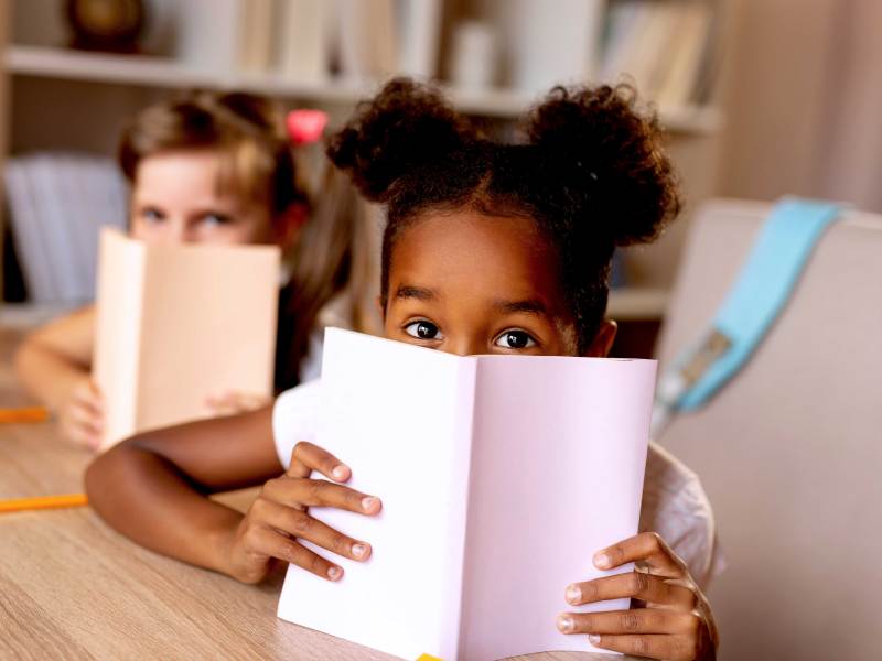 Zwei Mädchen verstecken sich hinter ihren Büchern, die sie in der Bibliothek lesen.