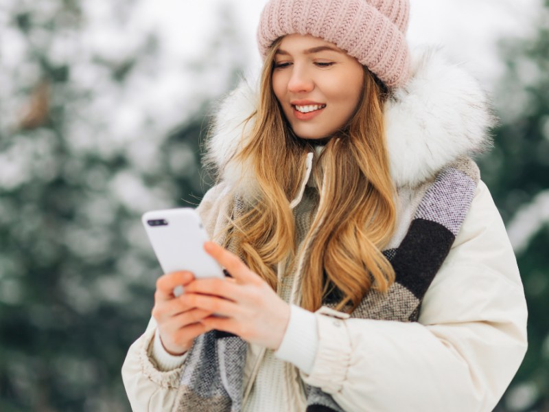 Eine Frau in Winterjacke und Mütze steht in einer Schneelandschaft und nutzt ihr Smartphone.