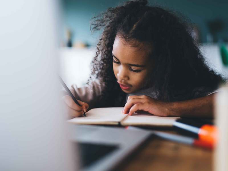 Ernsthaftes afroamerikanisches Mädchen mit lockigem Haar lernt und macht sich Notizen im Notizbuch, während es am Holztisch mit Laptop vor verschwommenem Hintergrund im Wohnzimmer sitzt