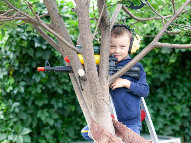 Kleiner Junge sitzt mit Ohrenschützern und einem Spielzeuggewehr in einem Baum und schaut verschmitzt in die Kamera.