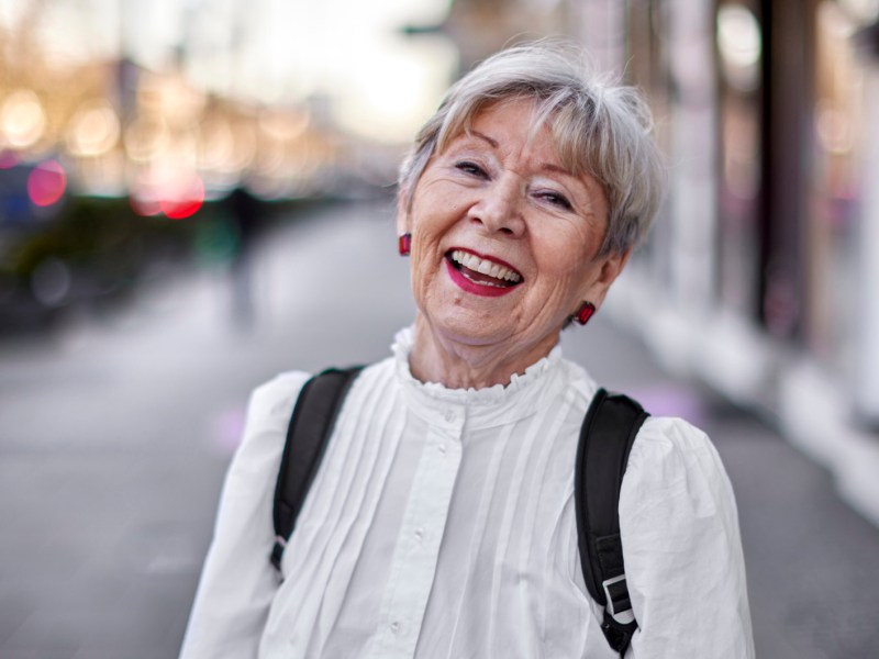 Modern gekleidete, ältere Frau mit einer weißen Bluse, roten Ohrringen und weißen Haaren lacht in die Kamera.